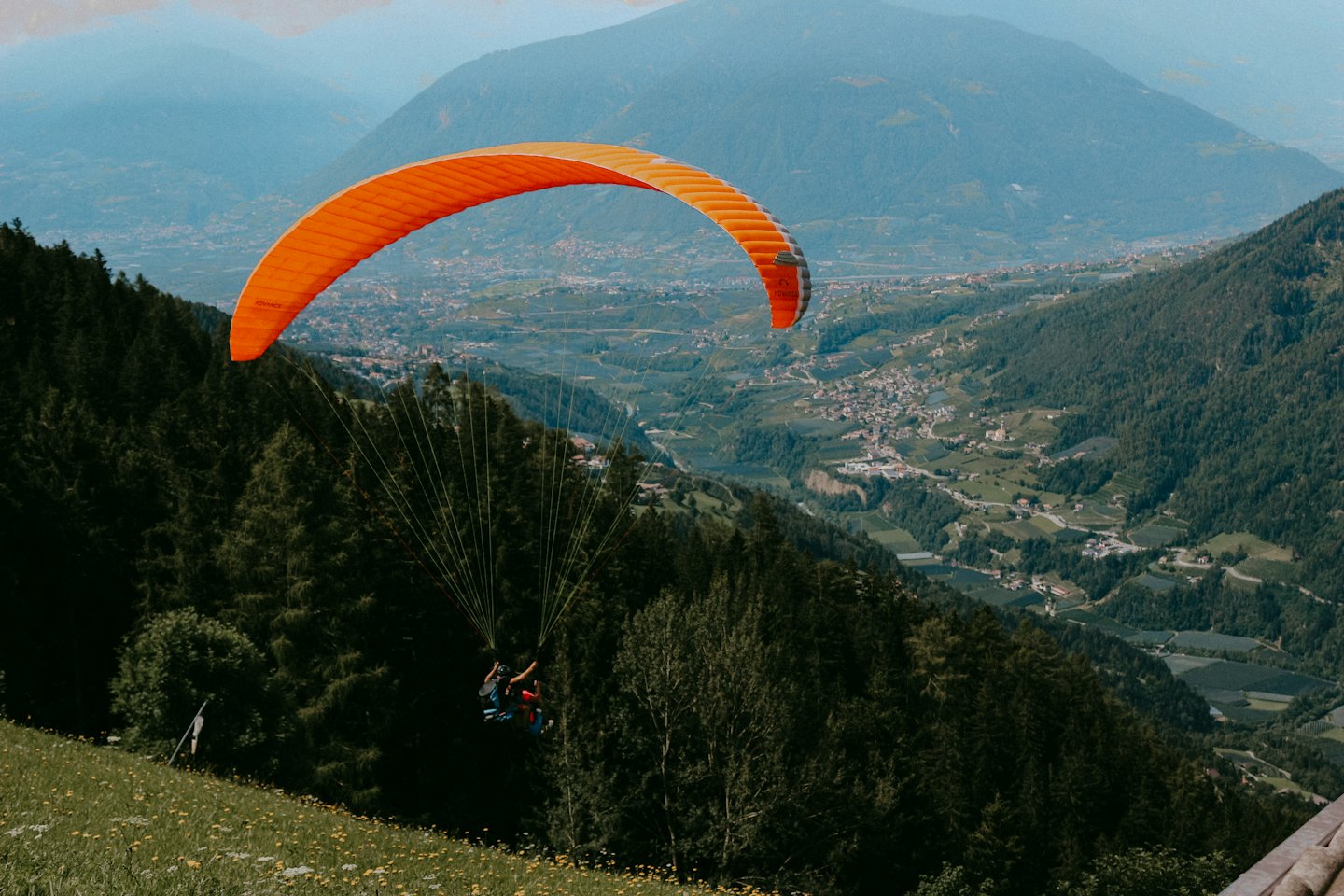 Ein orangefarbener Gleitschirm fliegt über ein grünes Gebirgstal.