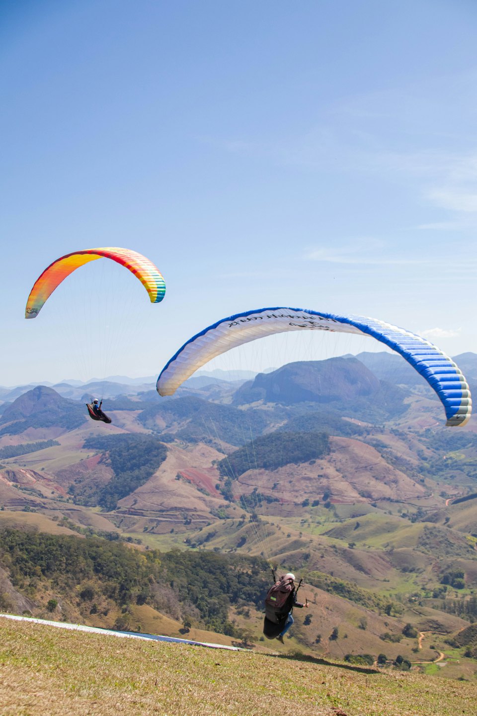 Gleitschirmflieger gleiten über grüne und braune Berge unter blauem Himmel.