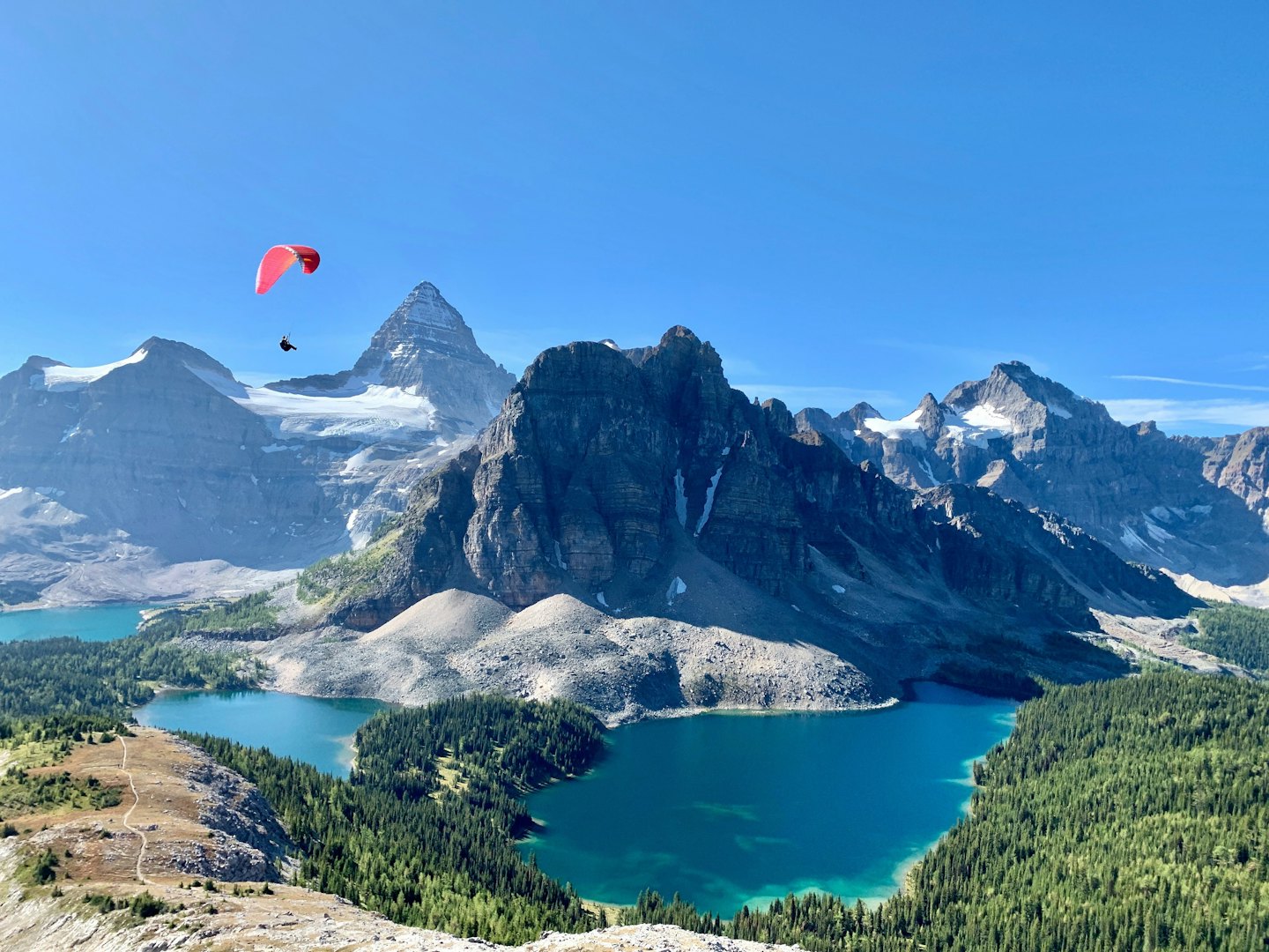 Ein Gleitschirmflieger fliegt über schneebedeckte Berge und türkisfarbene Alpenseen.