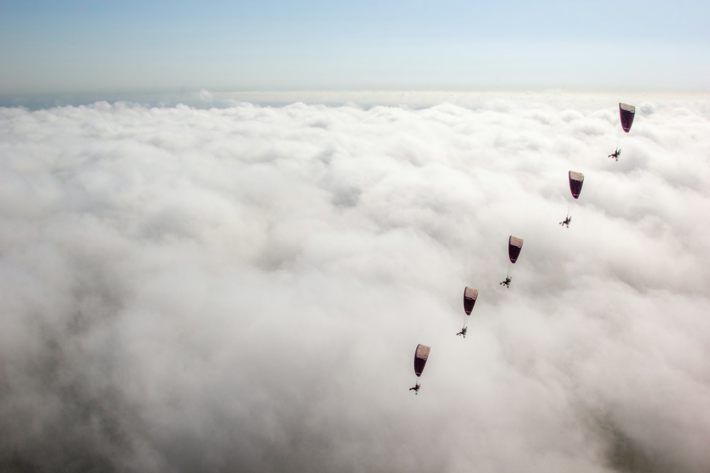 Fünf lila Gleitschirmflieger fliegen in Formation über dichten weißen Wolken.