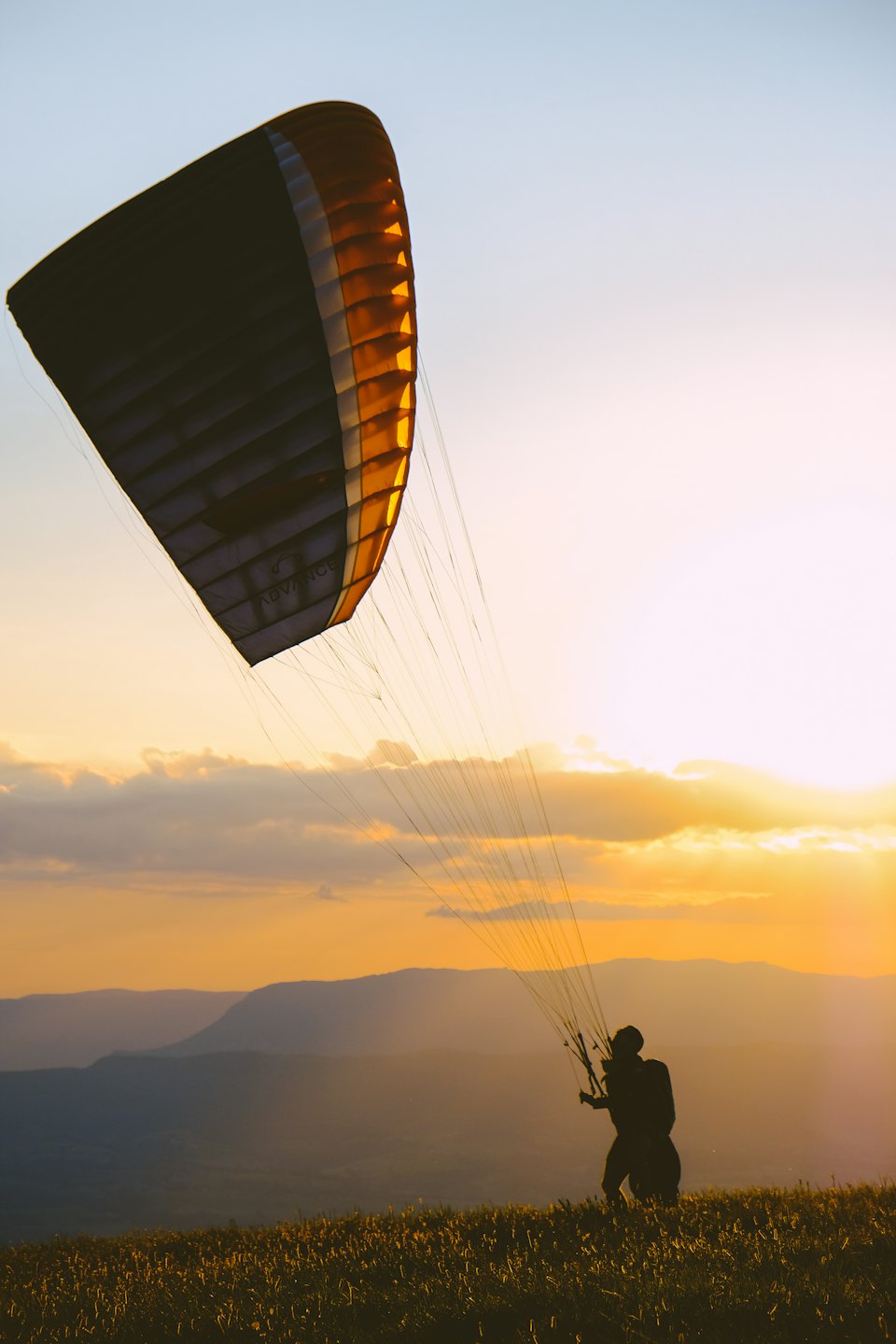 Gleitschirm mit entfalteter Tragfläche auf einem Berggipfel bei Sonnenuntergang.
